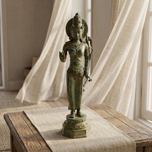 Può includere: Una statua color bronzo di una divinità è posta su un tavolo di legno. La statua presenta dettagli intricati, tra cui un'aureola e abiti ornati. La statua è alta circa 30 cm. La stanza è illuminata dalla luce naturale.