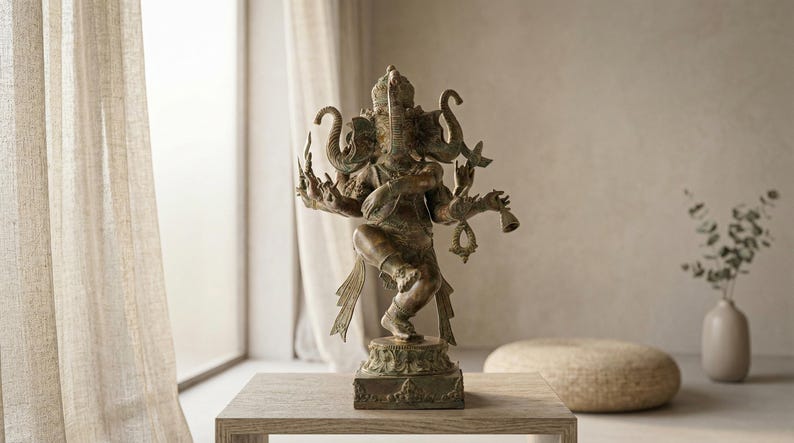 Op de afbeelding: Bronzen beeld van de hindoe&iuml;stische godheid Ganesha, met een olifantenkop en meerdere armen. Het beeld staat op een houten tafel. Een vaas met groen en een ronde poef zijn ook in de sc&egrave;ne, bij een raam.