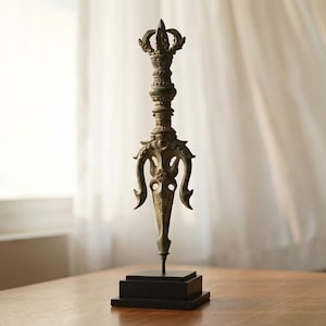 Puede incluir: Un vajra de metal ornamentado, de color bronce, un objeto ritual, se encuentra sobre una base negra. El vajra presenta detalles intrincados y una parte superior en forma de corona. Se exhibe sobre una mesa de madera frente a una ventana con cortinas blancas transparentes.