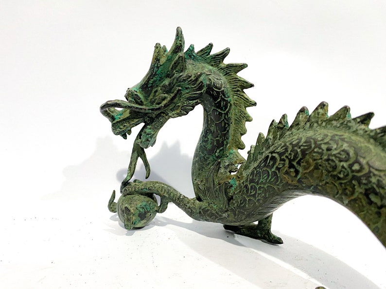 May include: A detailed bronze dragon sculpture with a verdigris finish. The dragon is holding a sphere in its claw and has a detailed, textured body. The sculpture is on a white background.