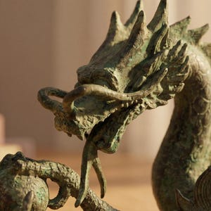 May include: A detailed bronze dragon sculpture with a textured surface. The dragon is depicted with an open mouth, sharp teeth, and intricate scales. The sculpture is a decorative item, possibly for home decor.
