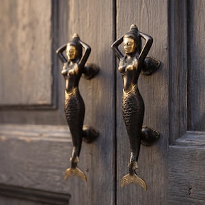 May include: Two bronze mermaid door handles are mounted on a weathered wooden door. The mermaids have their arms raised above their heads. The door handles have a gold finish on the face and tail.