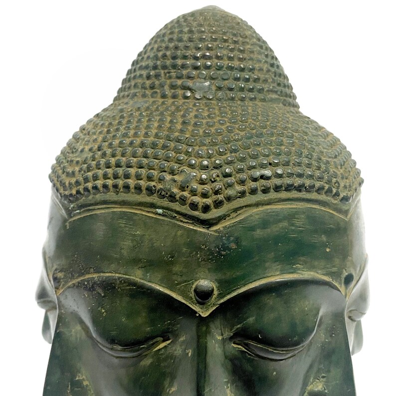 May include: A close-up of a weathered, green-toned Buddha head statue. The statue features closed eyes, a serene expression, and a detailed, textured hairstyle. The head is set against a plain white background, highlighting the sculpture's intricate details.