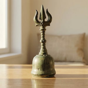 Peut inclure: Cloche en bronze avec un sommet en forme de trident, posée sur une table en bois. La cloche présente un design orné et une base évasée. L'arrière-plan comprend une fenêtre, un coussin et une plante en pot. La cloche mesure environ 15 cm de haut.
