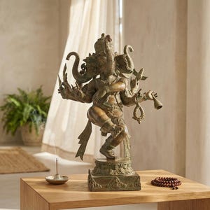 Op de afbeelding: Een bronskleurig metalen standbeeld van een godheid met meerdere armen en een olifantenkop, afgebeeld in een danshouding op een vierkante voet. De figuur houdt diverse symbolische voorwerpen vast en staat op een houten oppervlak.