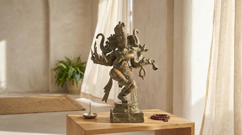 Op de afbeelding: Een bronskleurig metalen standbeeld van een godheid met meerdere armen en een olifantenkop, afgebeeld in een danshouding op een vierkante voet. De figuur houdt diverse symbolische voorwerpen vast en staat op een houten oppervlak.