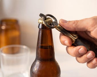 May include: A brown beer bottle being opened with a decorative bottle opener. The bottle opener has a dark handle with gold accents. The bottle is brown, and the cap is gold. Other jars and a glass are in the background.