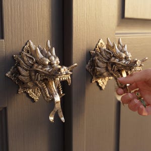 Könnte beinhalten: Zwei antike Türklopfer in Form von Drachenköpfen in Goldmetallic, montiert an einer dunkelgrauen Tür. Eine Hand greift nach einem Schlüssel im Schloss. Die Türklopfer sind sehr detailliert.