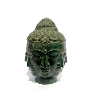 May include: A dark green, multi-faced Buddha head sculpture. The sculpture features four visible faces and a textured, rounded top. The faces have serene expressions and closed eyes. The sculpture is set against a white background.