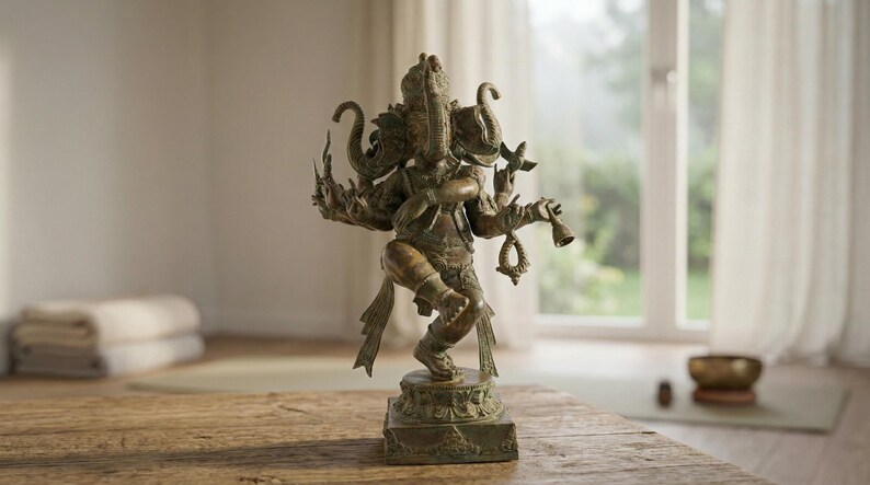Op de afbeelding: Bronzen beeld van de hindoe&iuml;stische godheid Ganesha, afgebeeld met een olifantenkop en meerdere armen, staand op een vierkante basis. Het beeld staat op een houten tafel, met een yogamat en opgevouwen handdoeken op de achtergrond.