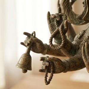 Op de afbeelding: Een bronzen standbeeld van een godheid met meerdere armen. De figuur houdt een kleine bel in de ene hand, een lusvormig touw in de andere en een kralenketting in een derde hand, terwijl een vierde hand in een open gebaar staat.