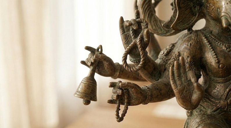 Op de afbeelding: Een bronzen standbeeld van een godheid met meerdere armen. De figuur houdt een kleine bel in de ene hand, een lusvormig touw in de andere en een kralenketting in een derde hand, terwijl een vierde hand in een open gebaar staat.