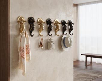 Könnte beinhalten: Eine Reihe von sieben seepferdchenförmigen Wandhaken in abwechselnd goldener und dunkelbronzener Ausführung. Die Haken sind an einer Wand montiert und halten ein gemustertes cremefarbenes Handtuch, einen Schlüsselanhänger aus Leder, einen kleinen Stoffbeutel und einen weißen Fedora-Hut.
