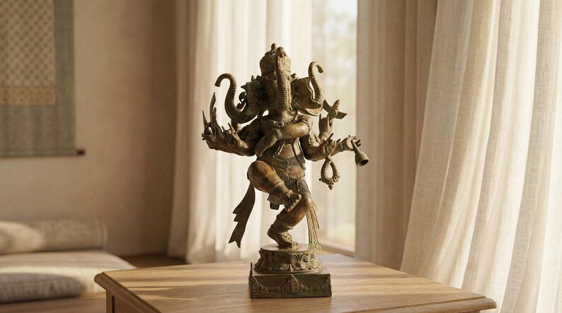 Op de afbeelding: Bronzen beeld van de hindoe-god Ganesha, met een olifantenkop en meerdere armen. Het beeld staat op een houten tafel, met een raam en transparante gordijnen op de achtergrond. Het beeld is ongeveer 30 cm hoog.