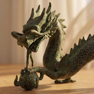 May include: A detailed bronze dragon sculpture with a textured, aged appearance. The dragon is posed with its head raised, holding a sphere. The sculpture features intricate scales and a spiky dorsal ridge, showcasing a mythical theme.