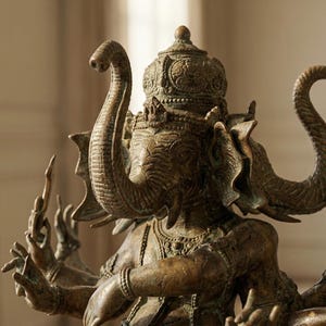 Op de afbeelding: Bronzen beeld van Ganesha, de hindoe&iuml;stische god van wijsheid en voorspoed. De sculptuur heeft een olifantenkop met meerdere armen, die verschillende objecten vasthouden. Het beeld is ingewikkeld gedetailleerd en heeft een verweerde uitstraling.