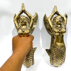 Könnte beinhalten: Zwei goldfarbene Meerjungfrauenstatuen mit ineinander verschränkten Händen zum Gebet. Die Meerjungfrauen sind mit langen, fließenden Haaren und Schuppen-besetzten Schwänzen dargestellt.