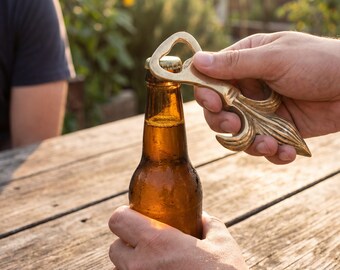 May include: A person opening a brown beer bottle with a decorative gold bottle opener. The bottle opener has a unique, ornate design. The scene is set outdoors on a wooden table.