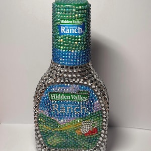 May include: A Hidden Valley The Original Ranch bottle completely covered in small, sparkling rhinestones. The neck features green and blue crystals, while the body is adorned with silver, green, and multi-colored stones forming a landscape scene.