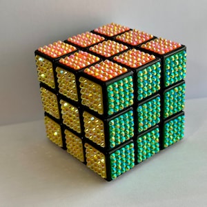 May include: A bedazzled Rubik's Cube with a black frame. The cube's faces are covered in iridescent rhinestones in shades of yellow, orange, and teal. The cube is a classic puzzle toy.