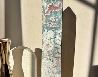 Op de afbeelding: Een hoge, gepolijste kristallen toren met een zeshoekige punt, voorzien van een gemarmerd patroon van mosgroen, crème, wit en roodbruine insluitingen door de steen, rechtopstaand op een getextureerde, cilindrische stenen voet.