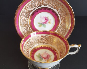 May include: A vintage teacup and saucer set with a pink and gold floral design. The cup and saucer have a white background with a pink rim and gold accents. The floral design features a pink rose with gold leaves.