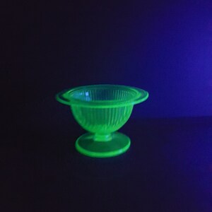 Puede incluir: Un cuenco de vidrio verde con un diseño acanalado y una base de pedestal. El cuenco está iluminado con una luz negra, lo que hace que brille.