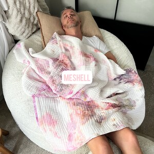 May include: A person is sleeping on a large, white, round beanbag chair. They are covered with a white, pink, and orange tie-dye blanket. The blanket has the word "MESHELL" printed on it.