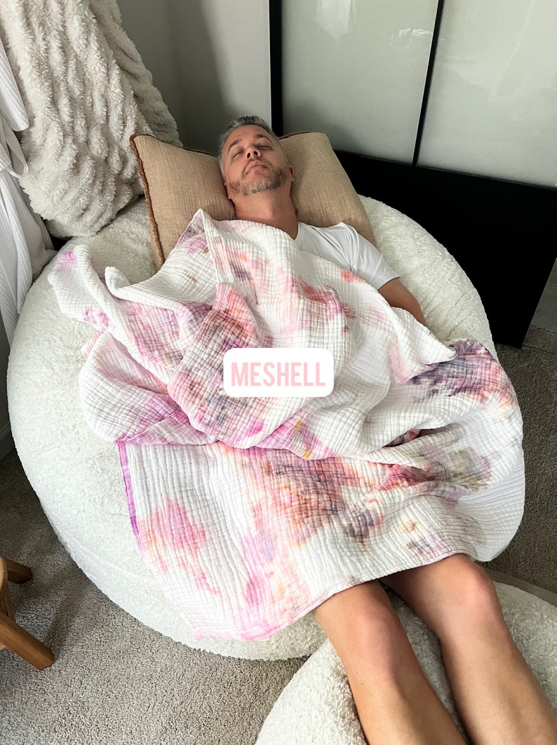 May include: A person is sleeping on a large, white, round beanbag chair. They are covered with a white, pink, and orange tie-dye blanket. The blanket has the word "MESHELL" printed on it.