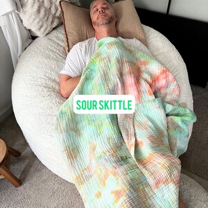 May include: A person is lying on a large, white, fluffy bean bag chair. They are covered with a green, orange, and pink tie-dye blanket. The blanket has the words "Sour Skittle" printed on it.