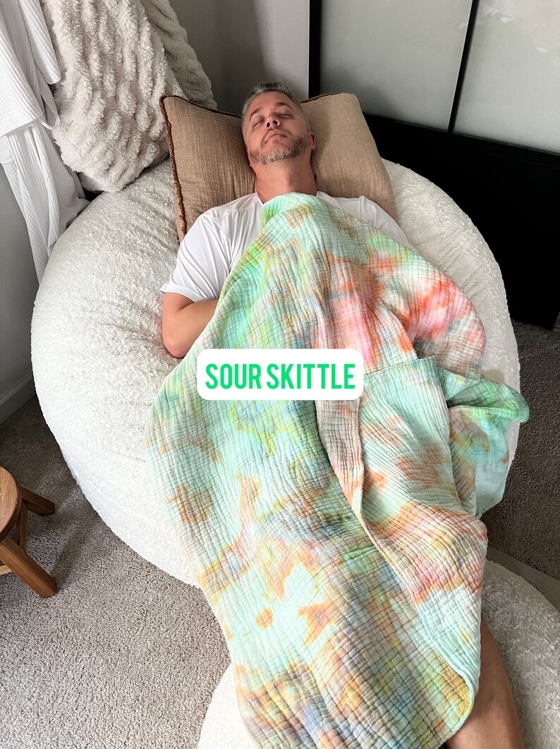 May include: A person is lying on a large, white, fluffy bean bag chair. They are covered with a green, orange, and pink tie-dye blanket. The blanket has the words "Sour Skittle" printed on it.
