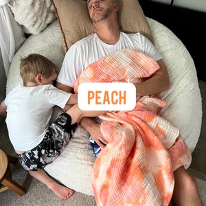May include: A father and son are napping on a large white beanbag chair. The father is wearing a white t-shirt and a peach-colored tie-dye blanket. The son is wearing a white t-shirt and black and gray shorts. The text "PEACH" is visible on the blanket.