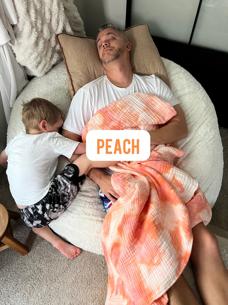 May include: A father and son are napping on a large white beanbag chair. The father is wearing a white t-shirt and a peach-colored tie-dye blanket. The son is wearing a white t-shirt and black and gray shorts. The text "PEACH" is visible on the blanket.