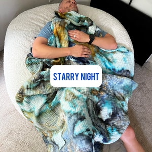 May include: A person is lying on a white beanbag chair, wrapped in a blue, green, and brown tie-dye blanket. The blanket is labeled "Starry Night".