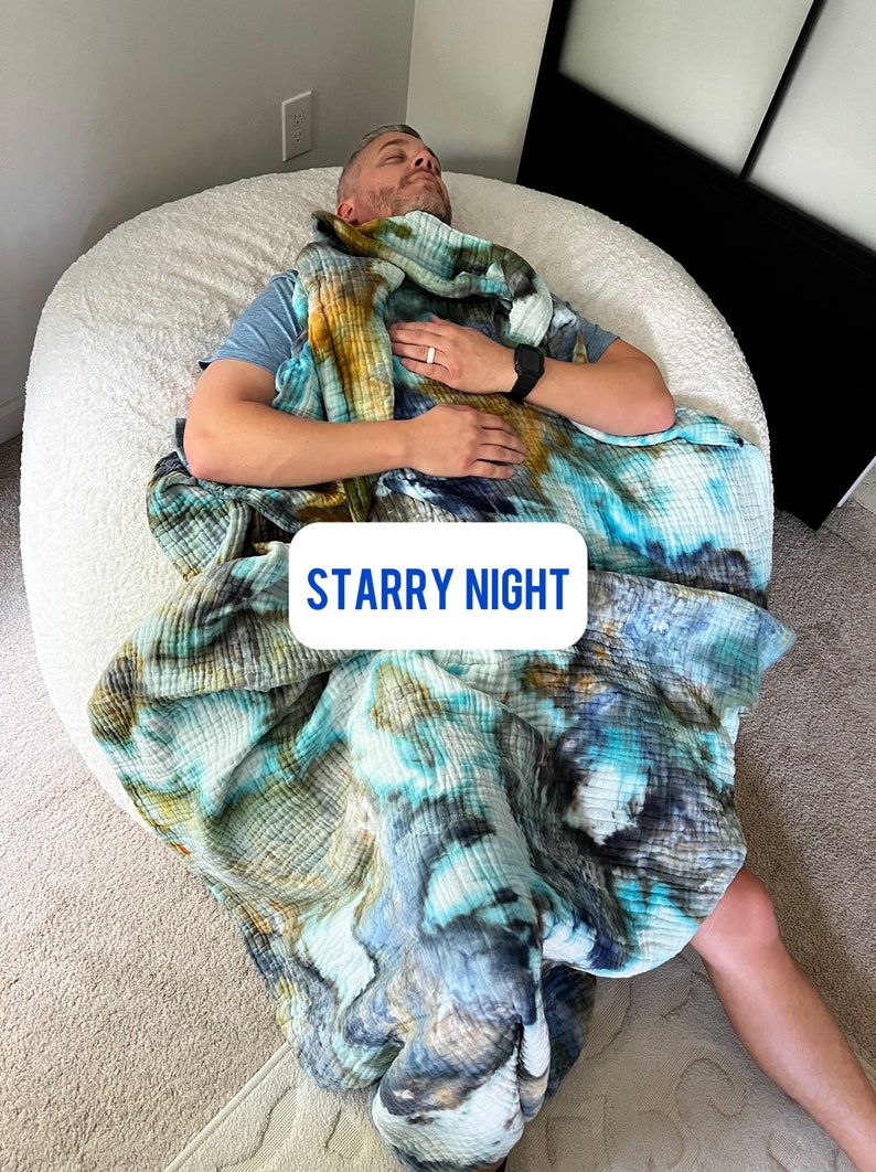 May include: A person is lying on a white beanbag chair, wrapped in a blue, green, and brown tie-dye blanket. The blanket is labeled "Starry Night".