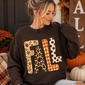 May include: A dark brown long-sleeve sweatshirt featuring the word FALL printed in large letters. Each letter has a distinct pattern: the F has a floral print, the A has a leopard print, the L has an orange gingham check, and the L has polka dots.