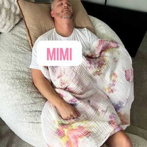 May include: A person sleeping in a white beanbag chair with a pink and white tie-dye blanket. The text "MIMI" is visible on the blanket.