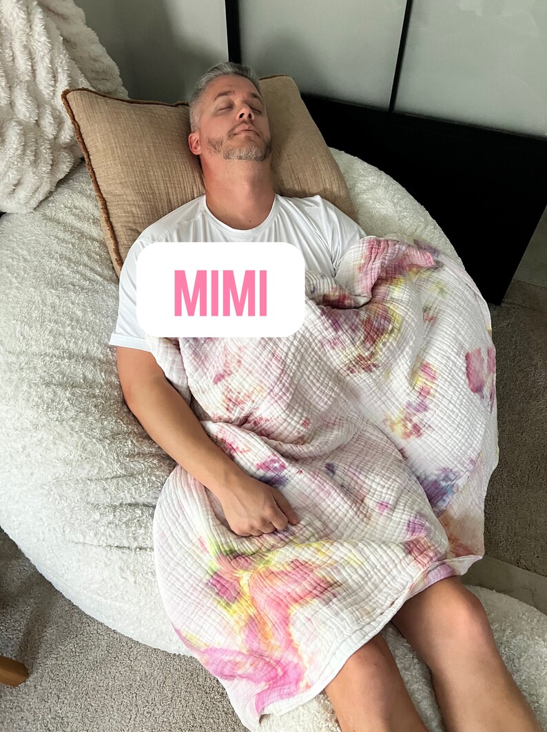 May include: A person sleeping in a white beanbag chair with a pink and white tie-dye blanket. The text "MIMI" is visible on the blanket.