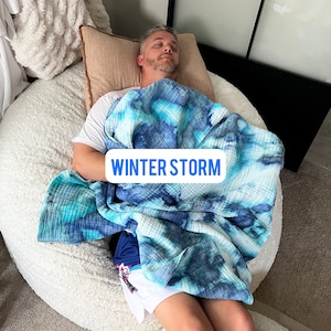May include: A person is lying on a large white beanbag chair, covered in a blue and white tie-dye blanket. The blanket has the text "WINTER STORM" printed on it.