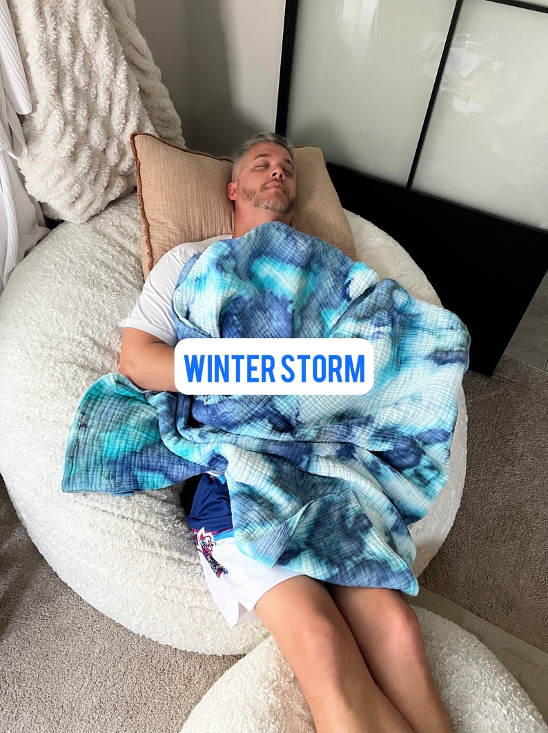 May include: A person is lying on a large white beanbag chair, covered in a blue and white tie-dye blanket. The blanket has the text "WINTER STORM" printed on it.