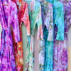 May include: Five tie-dye robes hanging on a white rack. The robes are in various shades of purple, pink, green, blue, and orange.