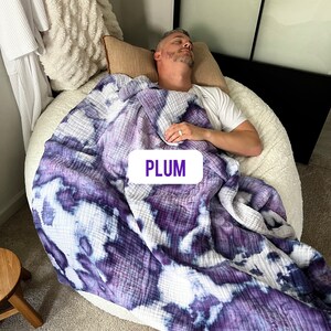 May include: A person is sleeping on a white bean bag chair covered with a purple and white tie-dye quilted blanket. The blanket has the word "PLUM" written on it in purple.