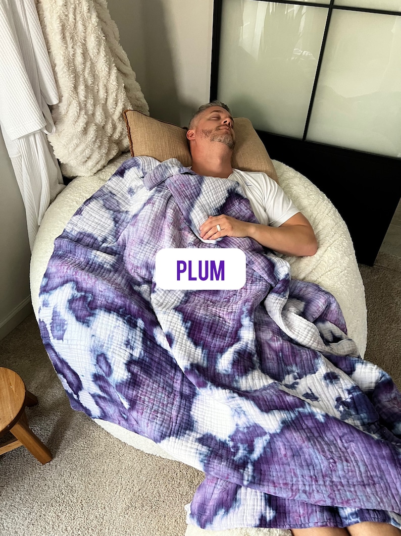 May include: A person is sleeping on a white bean bag chair covered with a purple and white tie-dye quilted blanket. The blanket has the word "PLUM" written on it in purple.