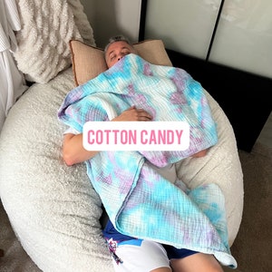 May include: A person is lying on a large, white, fluffy beanbag chair. They are covered with a blue and purple tie-dye blanket. The blanket has the words "COTTON CANDY" printed on it.