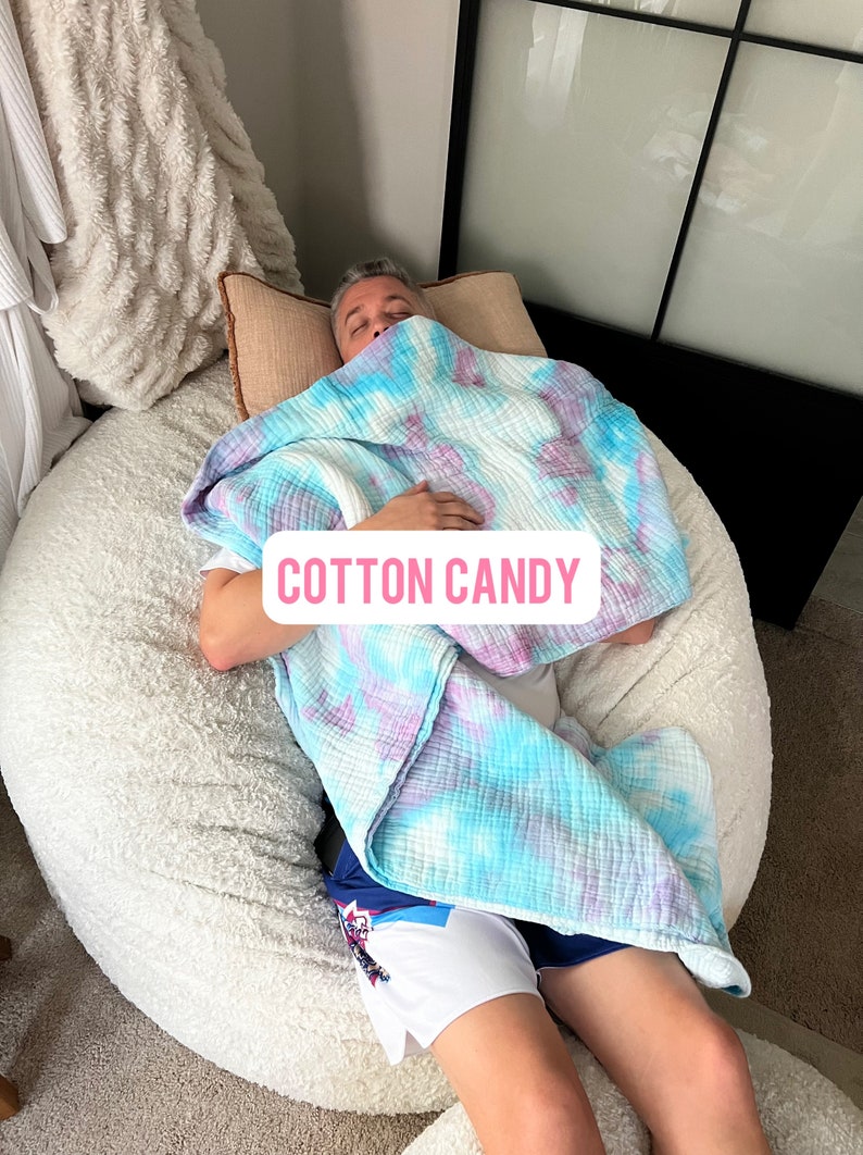 May include: A person is lying on a large, white, fluffy beanbag chair. They are covered with a blue and purple tie-dye blanket. The blanket has the words "COTTON CANDY" printed on it.