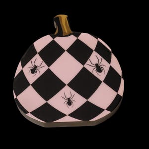 May include: A decorative pumpkin with a black and pink checkered pattern and three black spiders. The pumpkin has a gold stem and is set against a black background. Perfect for Halloween.