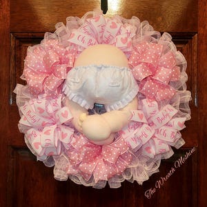 May include: A pink and white baby shower wreath with a baby doll wearing a white diaper and pink polka dot skirt. The wreath is decorated with pink and white ribbons and bows, and the text "Oh Baby!" is printed on the ribbons.