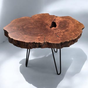May include: A live edge coffee table with a natural wood grain finish. The table has an organic shape with a dark hole in the centre. It is supported by three black hairpin legs, creating a modern, rustic aesthetic.