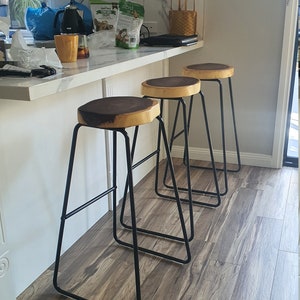May include: Three black metal bar stools with wooden seats. The stools have a triangular frame and are arranged in a row. The stools are in a kitchen with a white countertop and wood floor.