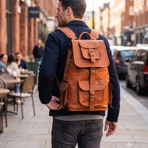 Könnte beinhalten: Ein beigefarbener Lederrucksack mit einer oberen Klappe mit Schnallenverschluss, einer Vordertasche mit Schnallenriemen und zwei Seitentaschen. Die Tasche verfügt über verstellbare Schultergurte, einen Tragegriff und messingfarbene Beschläge.